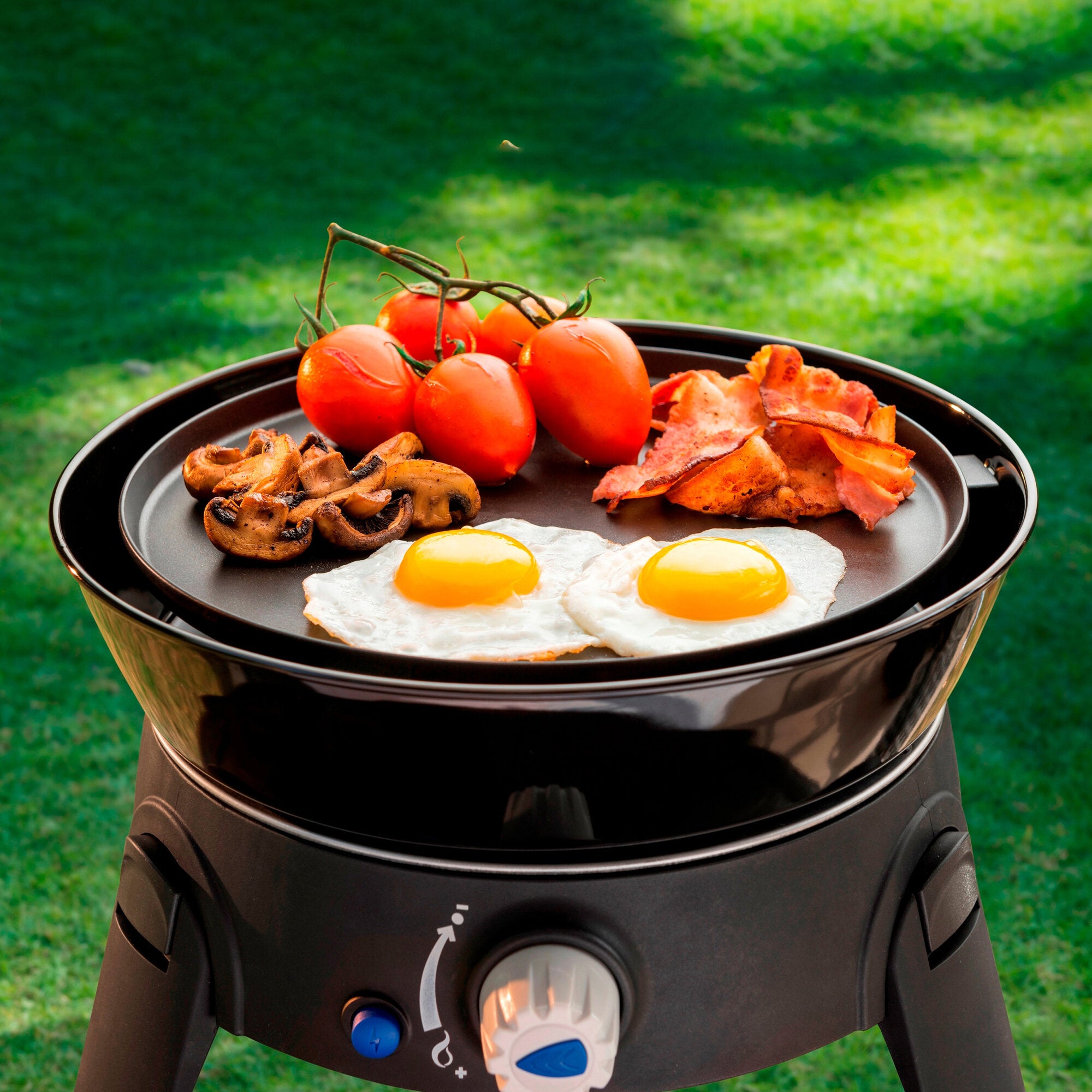 Runder Gasgrill im Freien, Deckel offen, Spiegeleier, Pilze und Tomaten am Zweig sowie Speck auf der Grillplatte, grüne Wiese im Hintergrund