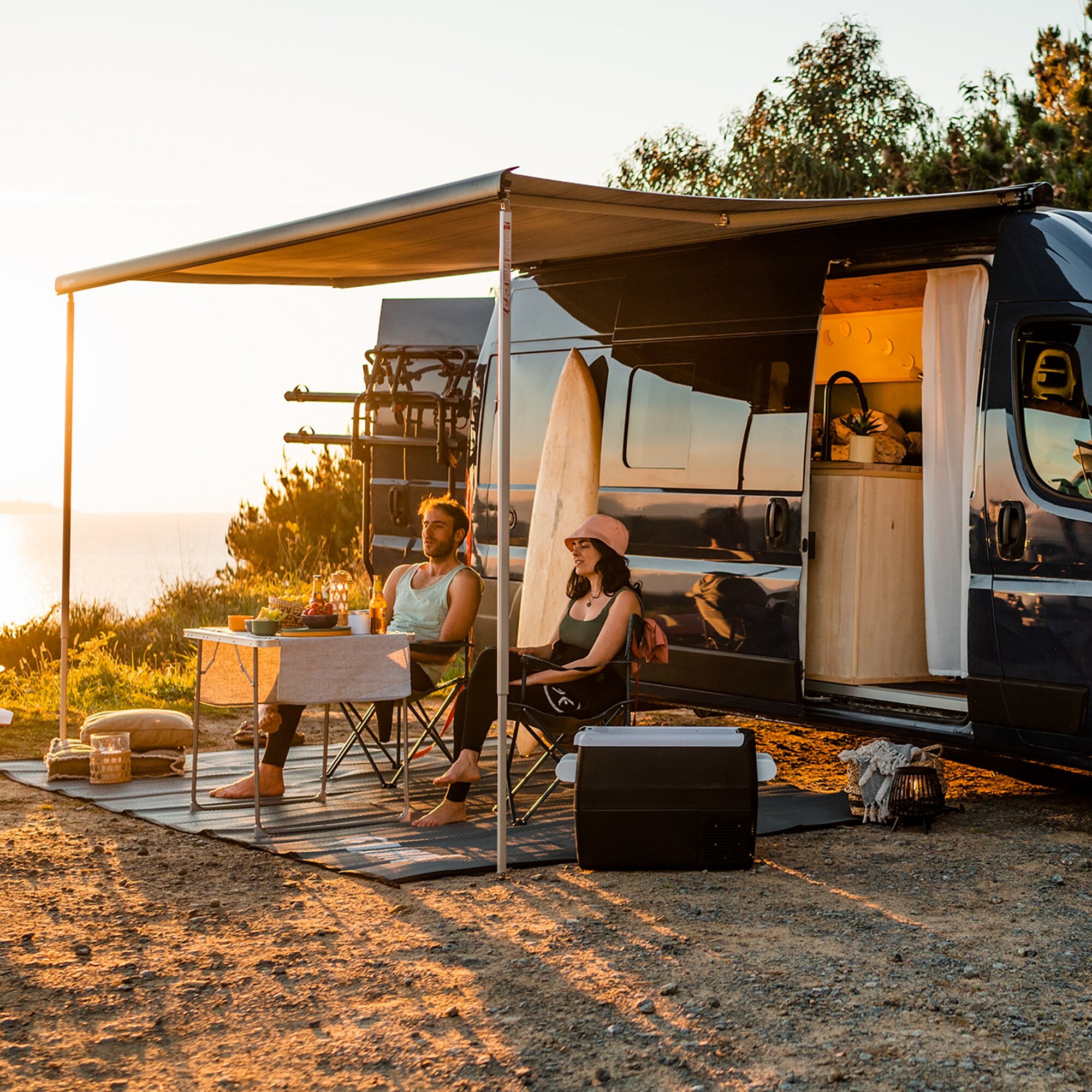 Seitenansicht eines Wohnmobils mit ausgefahrener Markise F80 Royal Grey, sonnendurchfluteter Campingplatz, Esstisch mit Gästen