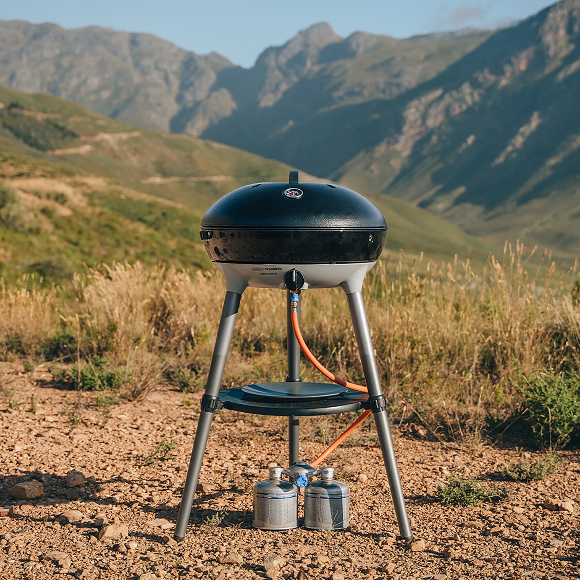 Gasgrill Kugelform auf drei Beinen mit schwarzem Deckel, orange Schlauch verbindet ihn mit zwei Propangasflaschen, felsiger Boden und Berglandschaft im Hintergrund