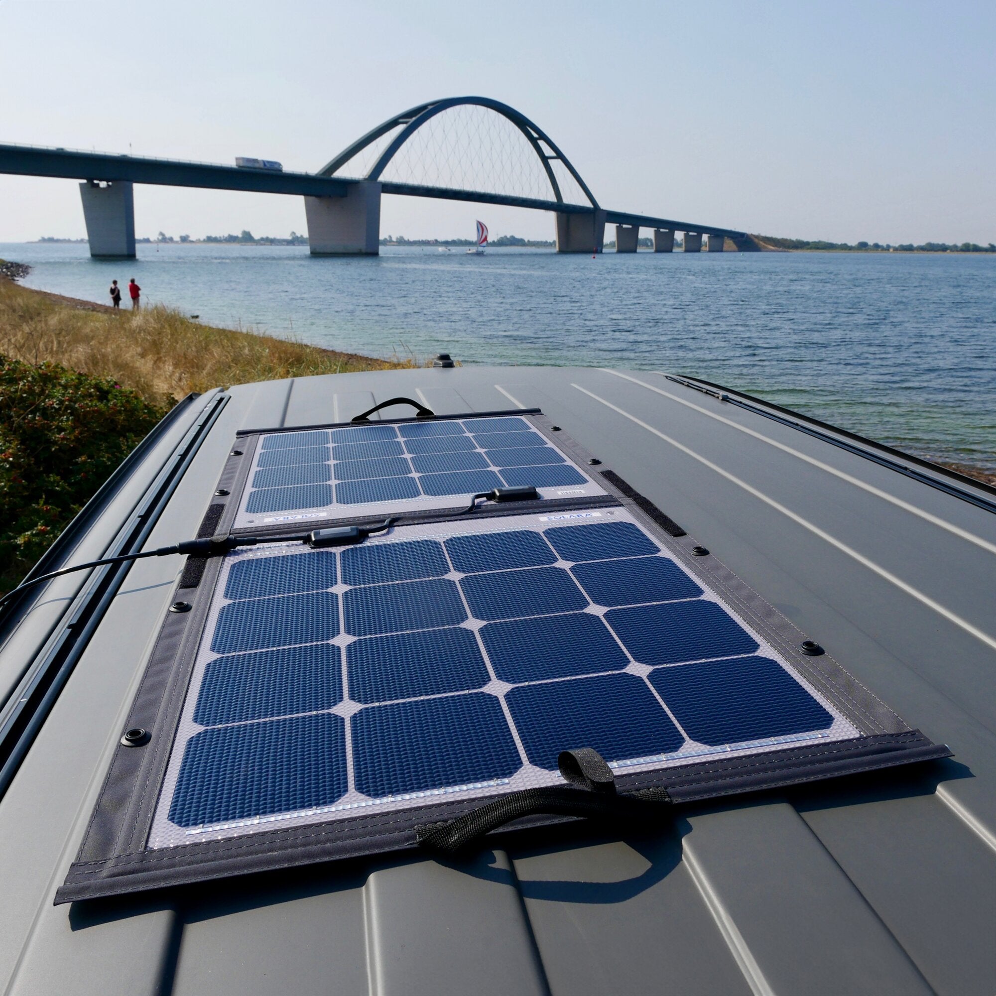 Auf dem Dach eines Campers liegt ein aufgeklapptes faltbares Solarmodul mit blauen Zellen, im Hintergrund eine Brücke über einem See, am Ufer zwei Personen