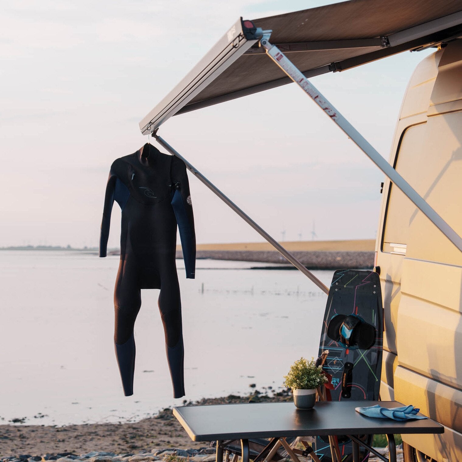Schwarzer Neoprenanzug hängt an einem Haken unter der Markise eines gelben Campers, daneben ein buntes Wakeboard und ein Tisch mit Pflanze und Handschuhen