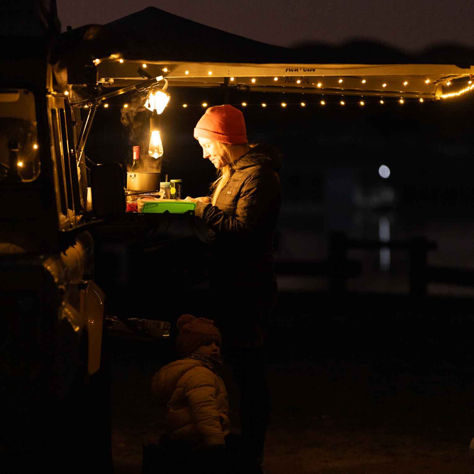 Nachtaufnahme in einem Camper: eine Person mit roter Mütze arbeitet an der Küchenplatte, von warmen Lichterketten beleuchtet; auf dem Tisch liegt ein grüner Silikonbehälter