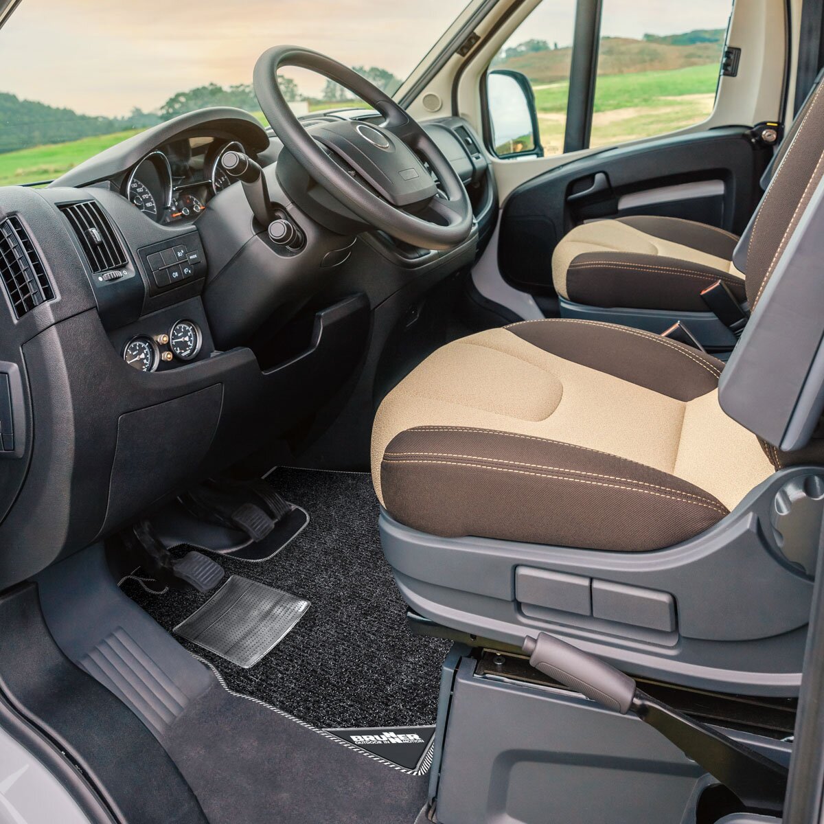 Innenraum eines Ford Transit mit Lenkrad und Pedalen; schwarze Teppich-Deluxe-Fußmatte im Vorderfußraum, Bezüge beige-braun