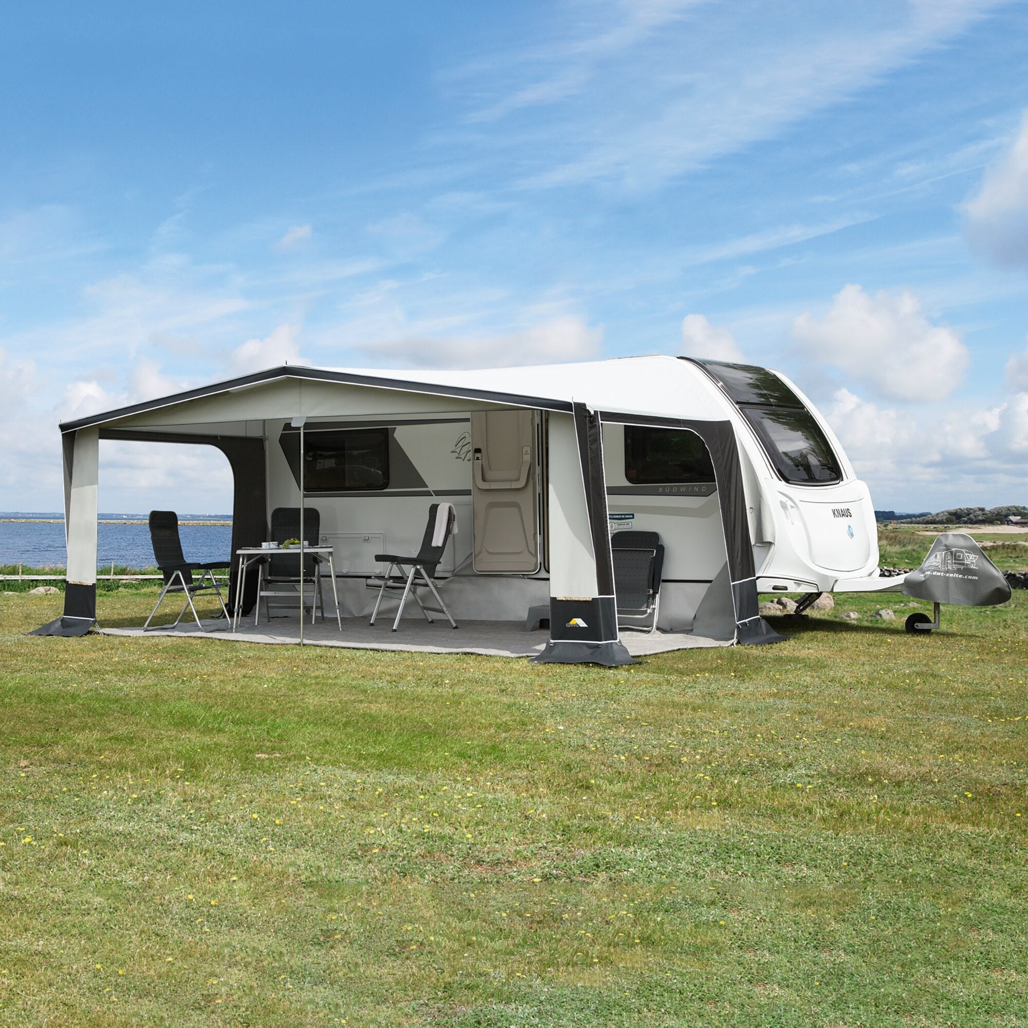 Großes Reisevorzelt am Wohnwagen, offene Sitzgruppe mit Klappstühlen und Tisch, Grasboden, Blick aufs Meer und blauen Himmel