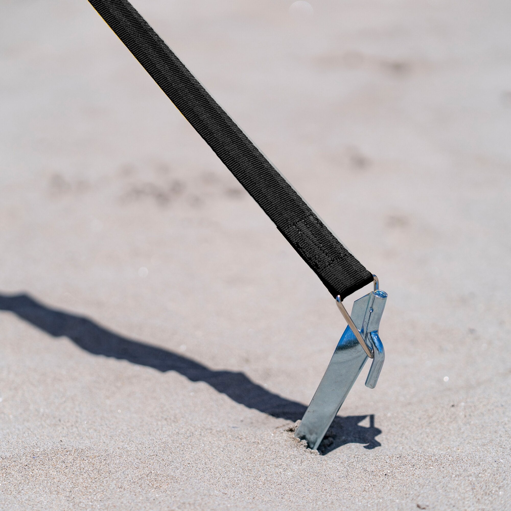 Schwarzes gewebtes Abspannband mit Metallhaken am unteren Ende, diagonal über Sandboden gespannt, an blauem Metallgestell befestigt