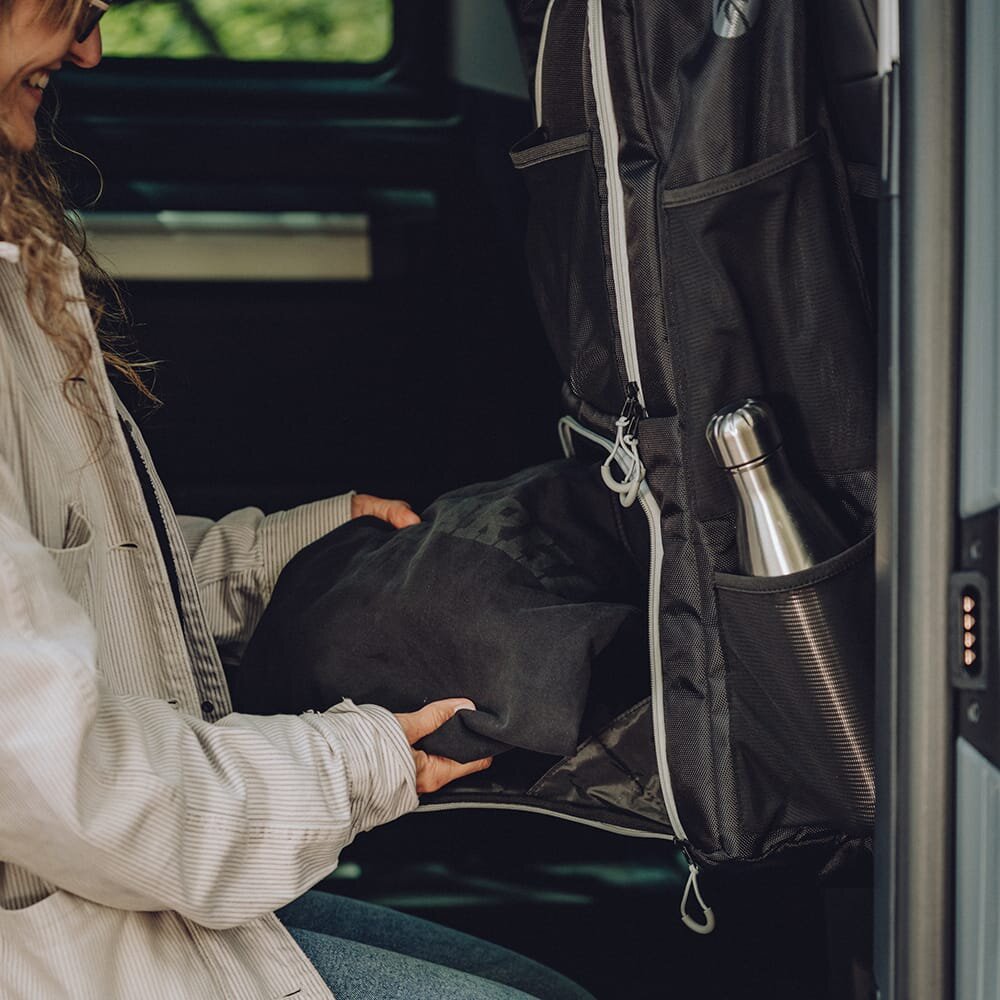 Schwarze SeatBOXX Organizer-Tasche hängt an der Rückenlehne eines VW T5/T6/T6.1; eine Person legt Decke hinein, im Seitfach steckt Wasserflasche