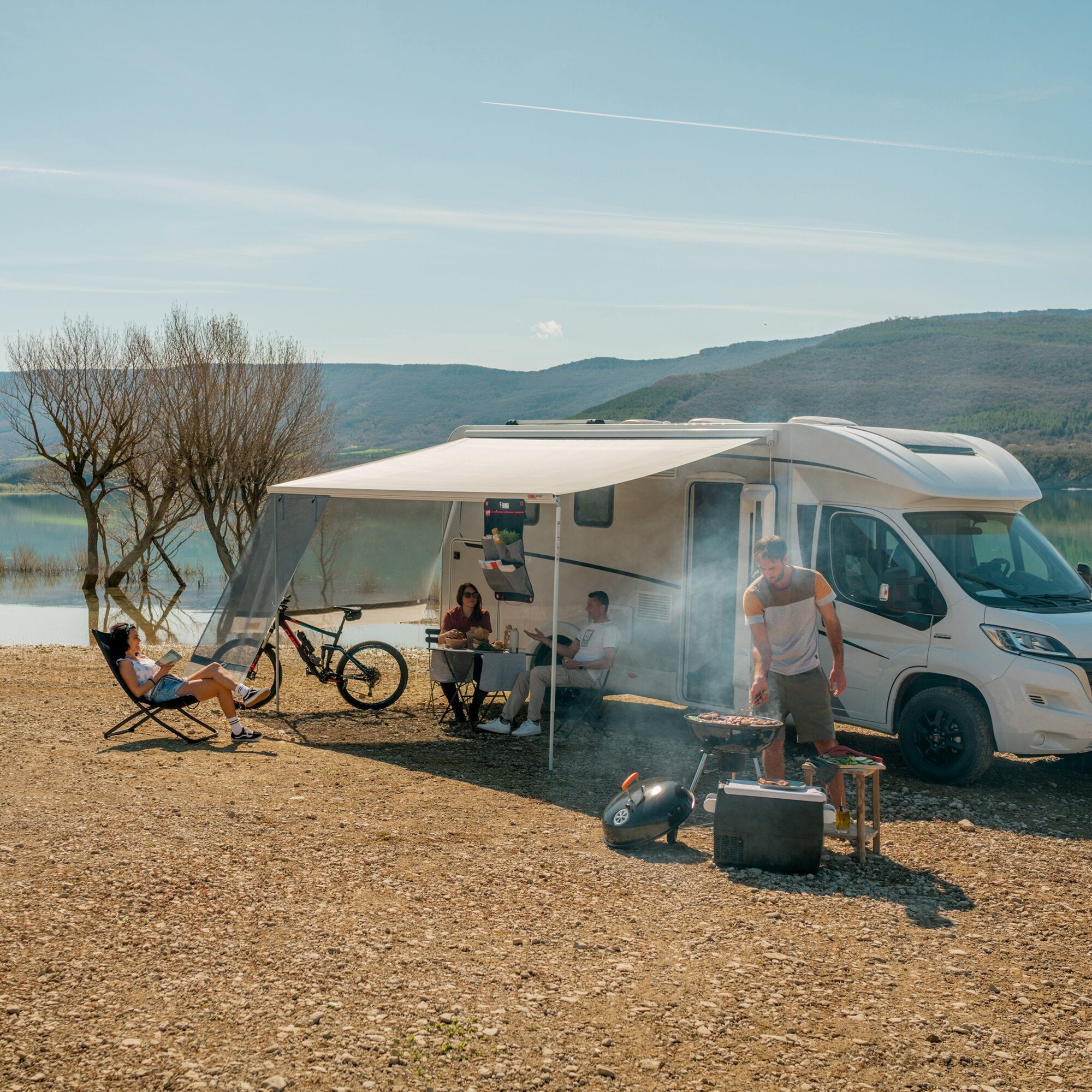 Wohnmobil mit ausgefahrener Seitenwand-Markise Sun View Side; draußen sitzen Personen am Tisch, daneben Fahrräder und ein Grill, Hügel im Hintergrund