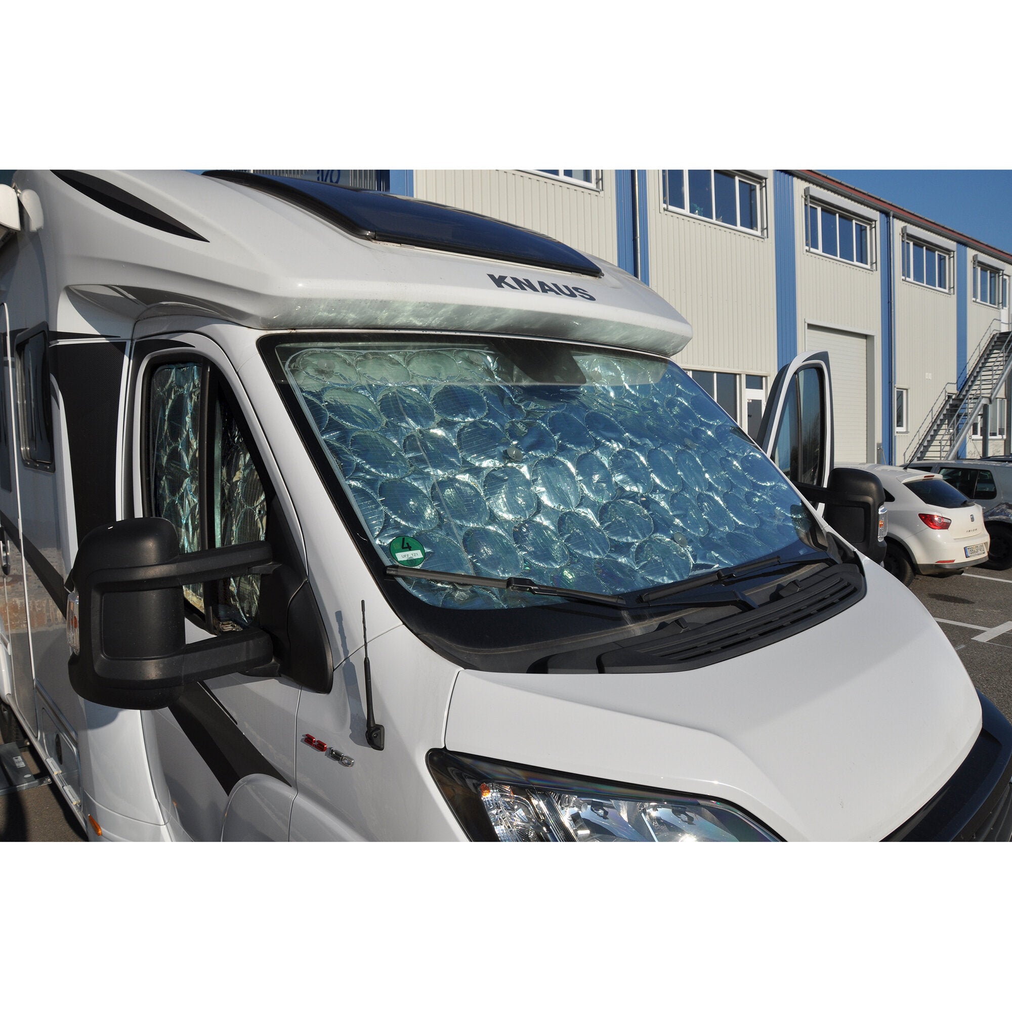 Weißes Wohnmobil mit reflektierender Windschutzscheiben-Wärmefolie, sichtbare Seitenspiegel und Frontpartie, Hintergrund Industriegebäude