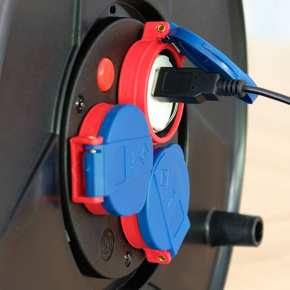 Detail eines USB-Steckdosenadapters mit rotem Rundrahmen und blauen Abdeckkappen; ein USB-A-Stecker steckt in einer weißen Buchse, schwarzes Kabel führt hinein