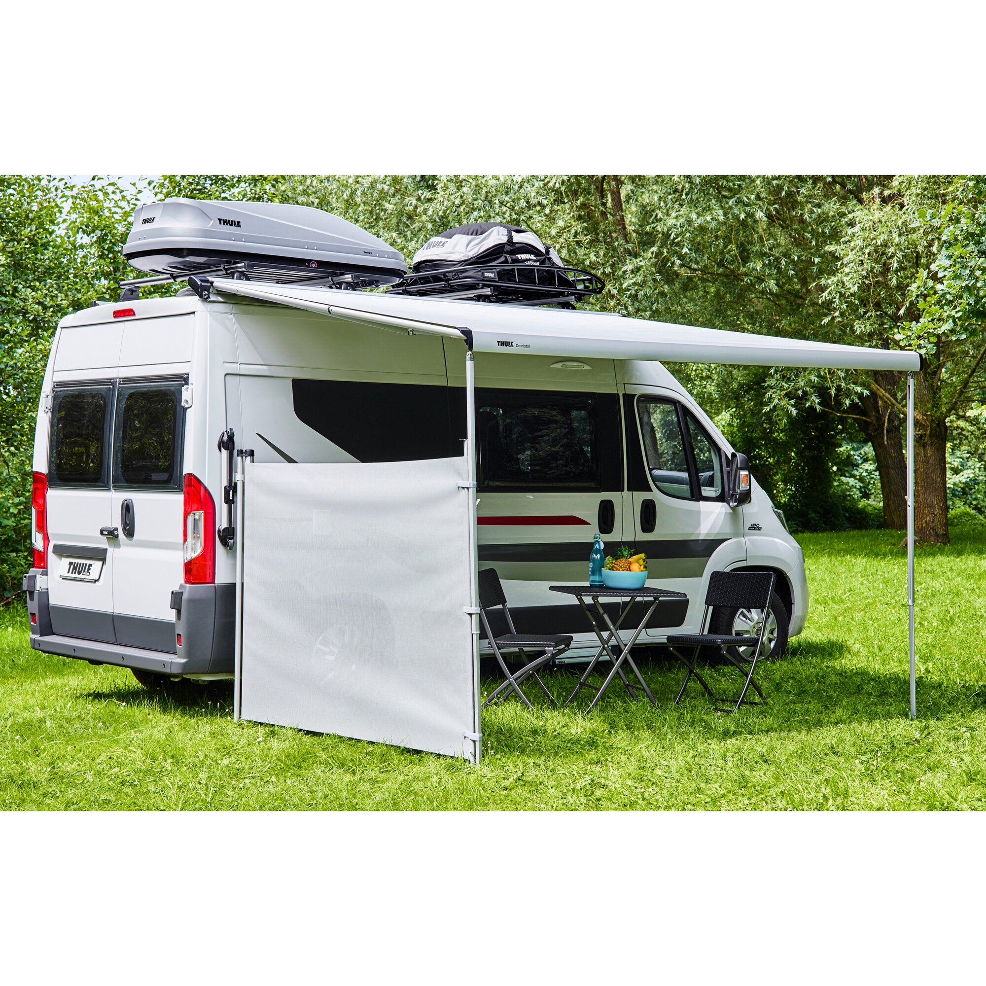 Ein weißer Campervan mit seitlichem Windschutz, davor ein Tisch und zwei Klappstühle; auf dem Dachträger graue Dachbox und schwarze Tasche