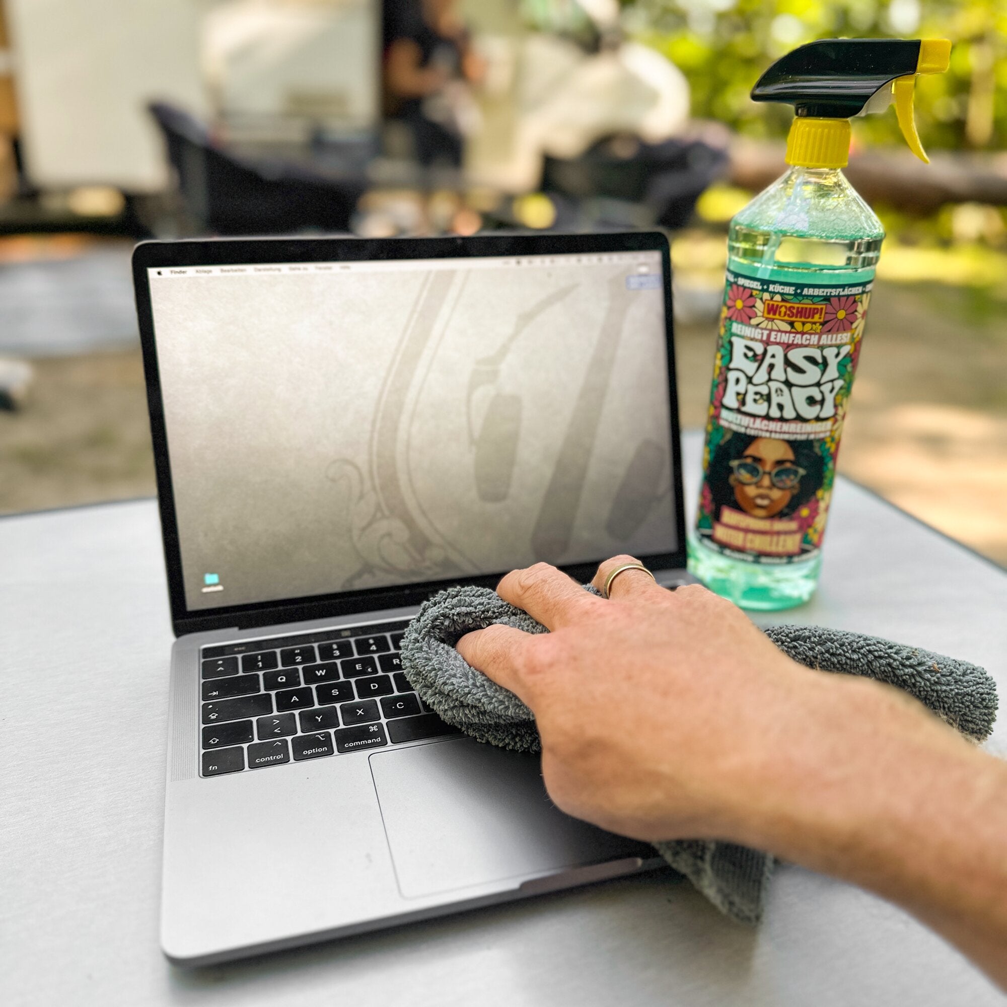 Eine Hand wischt mit Mikrofaser-Tuch über den Laptop, daneben steht eine Sprühflasche Reiniger mit gelbem Sprühkopf und floralem Etikett
