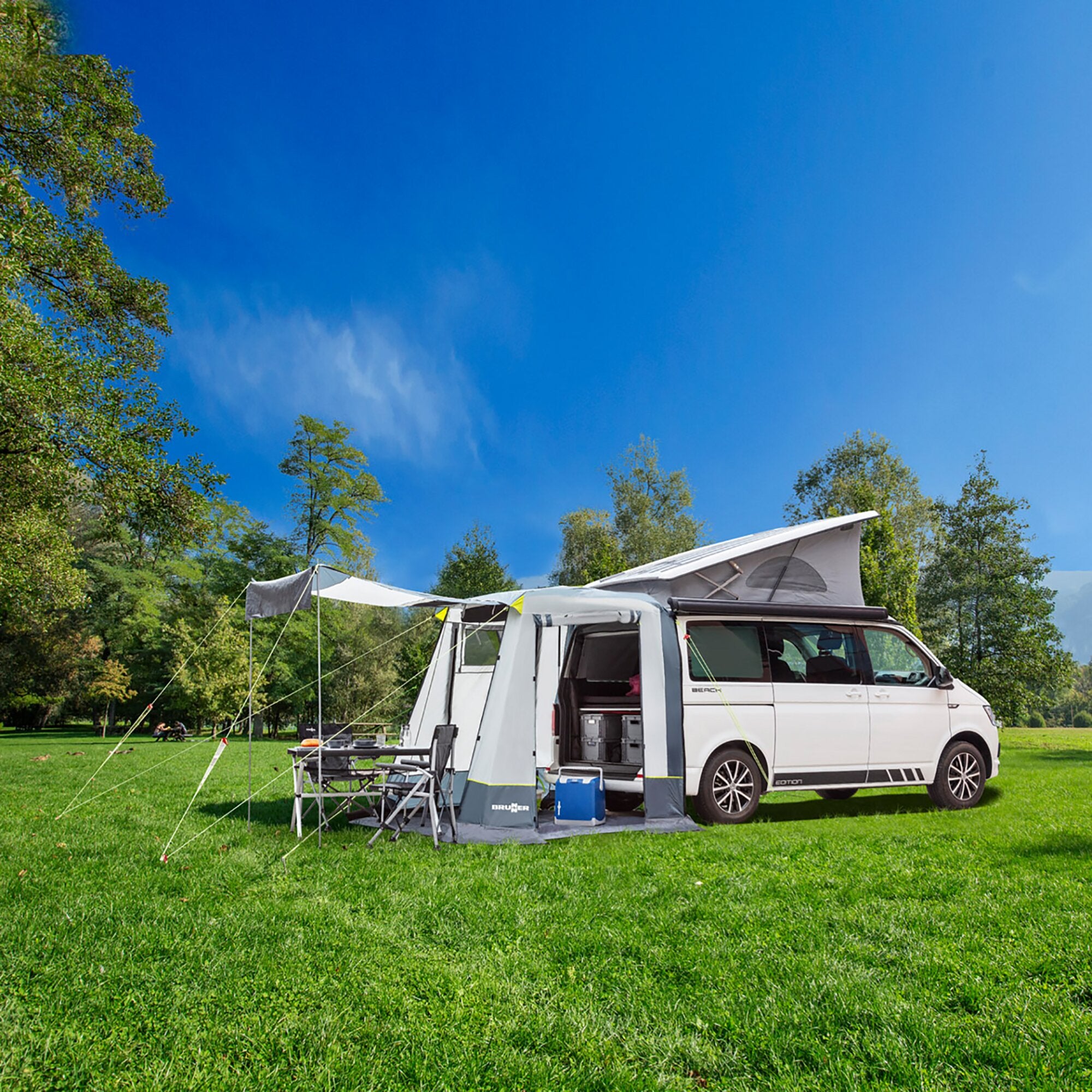 Weißer VW T5/T6 Camper mit Heckzelt Comet, aufgeklapptem Vorbau, Stühle, Tisch und Kühlbox auf grünem Rasen bei blauem Himmel