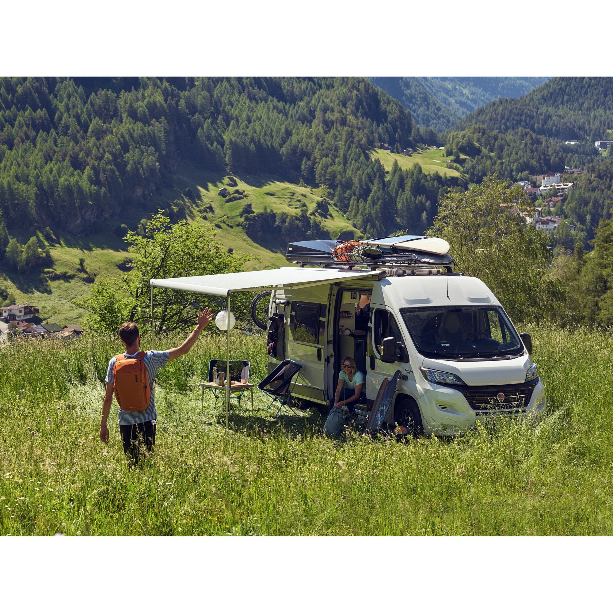 Weißer Camper mit seitlich ausgefahrener Markise, Dachgepäckträger, zwei Personen neben einem Tisch mit Getränken vor grünem Bergpanorama