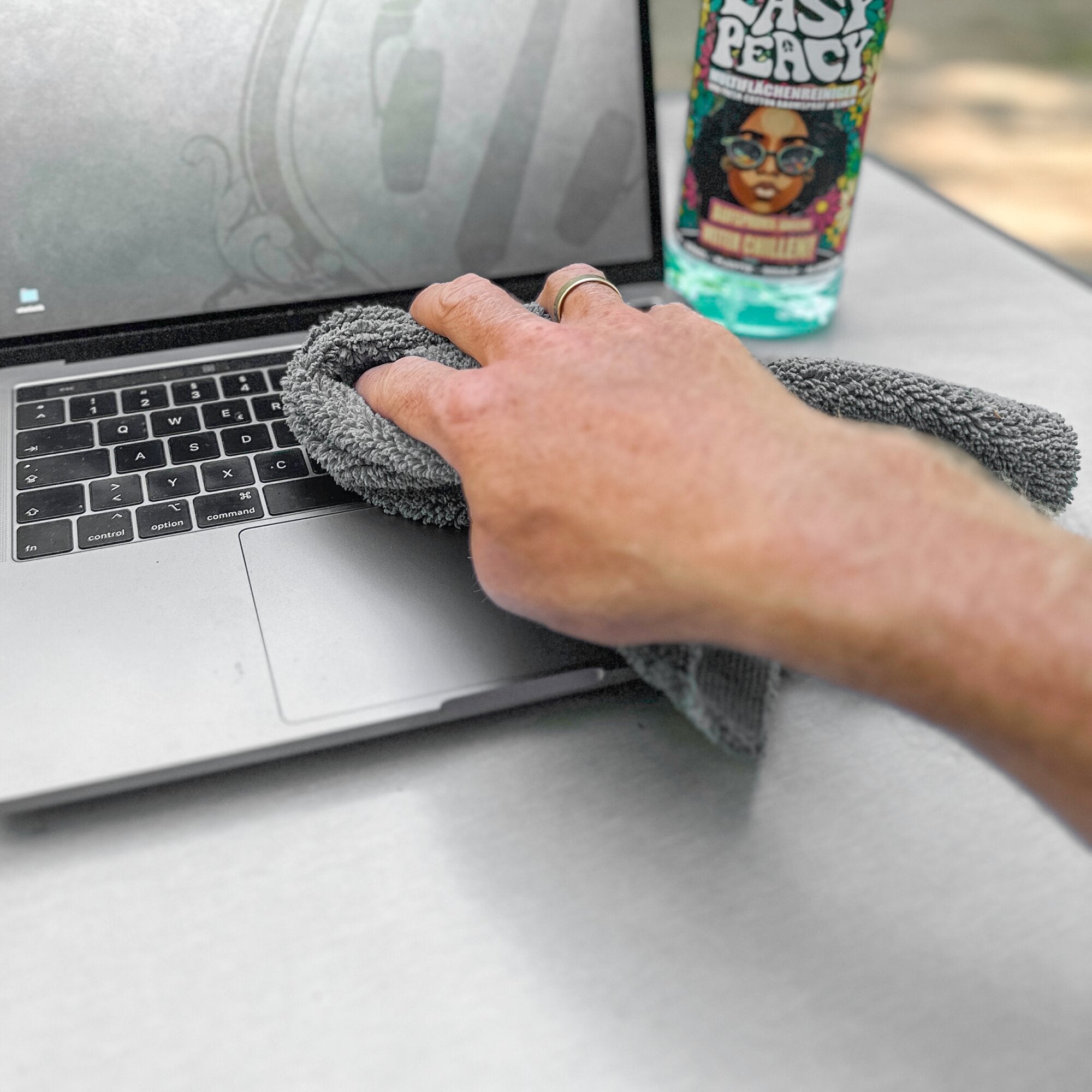 Eine Hand wischt mit einem grauen Hochflor-Reinigungstuch über den Laptop-Bildschirm, dahinter steht eine bunte Reinigungsflasche
