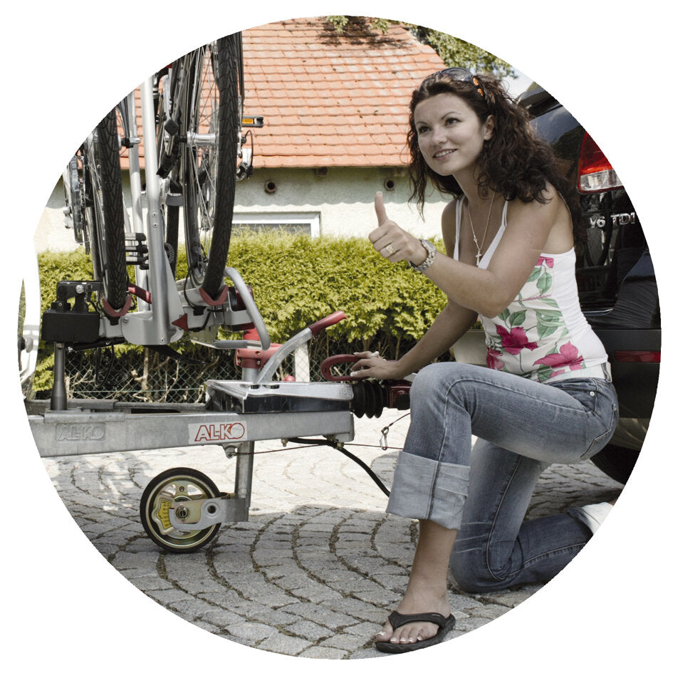 Frau kniet neben einem AL-KO Fahrradträger am Heck des Autos, der zwei Fahrräder transportiert; sichtbar ist ein Stützrad zur Stabilisierung