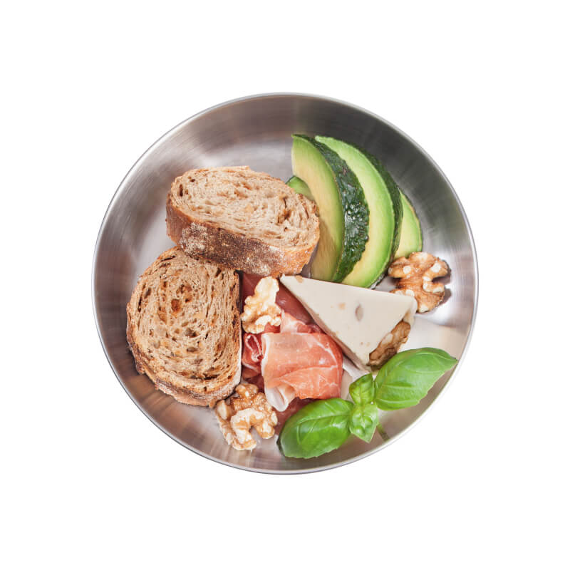 Runder Edelstahlteller mit Brot, Avocadoscheiben, Käse dreieck, Schinken, Walnüssen und frischen Basilikumblättern.