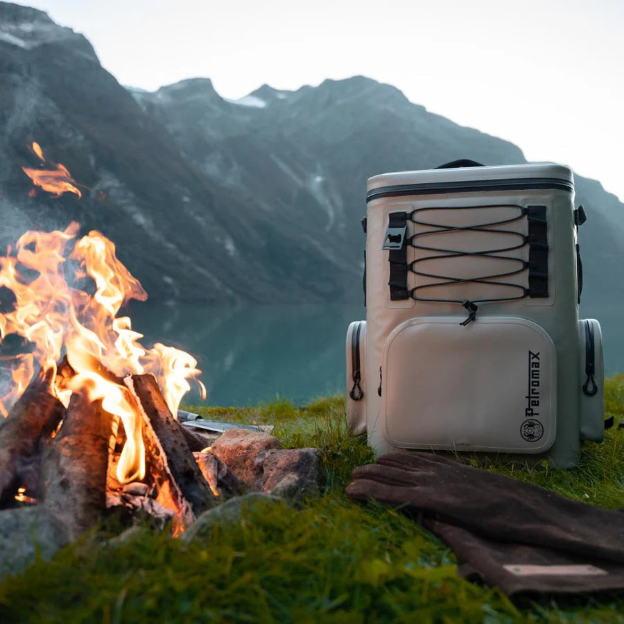 Beiger Petromax Kühlrucksack mit schwarzem Logo, seitlichen Griffen und elastischem Gummizug, stehend auf Gras nahe.